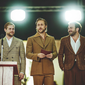 Mike Vogel, Ryan Gosling, and Derek Cianfrance at an awards ceremony. Wearing tailored suits, with subtle smiles. Dark stage, minimalistic design, bright spotlights.
