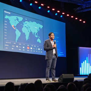 Parag Khanna, in a casual suit, speaking at a tech conference. The stage background is a large screen showing global economic data.
