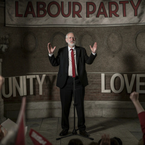 Jeremy Corbyn, dressed in a dark suit and red tie, addressing a rally. The backdrop features bold 'Labour Party' logos and messages of unity and love. Crowd is cheering.