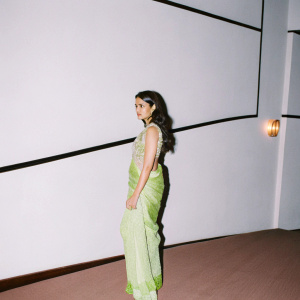 Lara Dutta, dressed in a vibrant green saree, poses elegantly on the set of a Bollywood movie premiere. The background is a minimalist, sleek design.