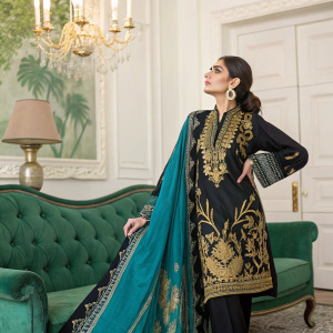 A woman modeling the Meraki Festival Collection by Ayeza Khan Wearing Lakhany. She's wearing a black and gold top with intricate designs and a flowing teal and black shawl. Background has an emerald green velvet sofa and a chandelier-like lamp.