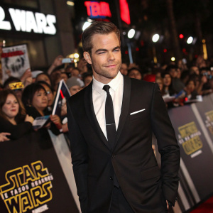Chris Pine at Comic-Con. Posing confidently in a black suit with crisp white shirt and black tie. Amidst sea of fans, Star Wars banners and props all around.
