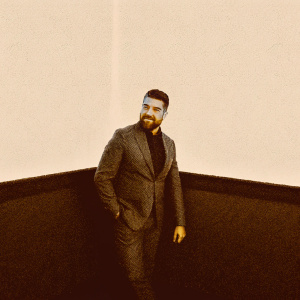 Josh Segarra, with a beard, dressed in a dark gray suit. Poses confidently in an empty, white room.