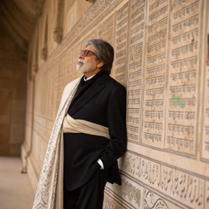 Renowned actor Amitabh Bachchan in a black suit with a formal shawl. Standing in front of a wall covered in ancient scriptures and intricate designs. Solemn expression.