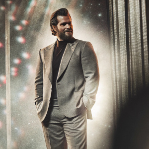 Henry Cavill, actor with chiseled jawline and rugged beard, standing solo at an event. He wears a stylish suit. Soft focus, colorful stage backdrop