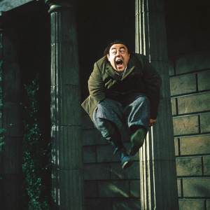 Close-up of Zero Mostel's face, mid-air pose. Comedic expression, wide-eyed and open-mouthed. Stone wall with vertical columns as backdrop.