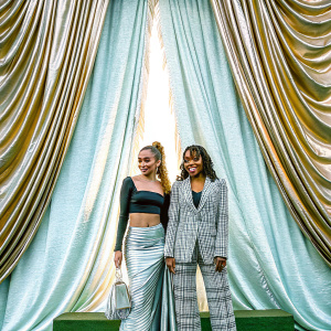 Brec Bassinger and Tetona Jackson at the Met Gala. Brec in a black crop top and a skirt with horizontal stripes. Tetona in a plaid-style jacket. Both standing in front of a gold and blue fabric backdrop.