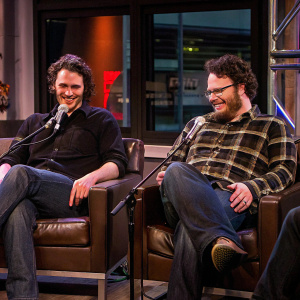 James Franco and Seth Rogen in casual outfits. Franco in black shirt, Rogen in plaid. Both seated in studio with SiriusXM logo backdrop. They are in midst of conversation, microphones in front of them, appear relaxed and engaged.