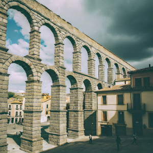 Historic Segovia aqueduct bridge stands in a bustling cityscape. Traditional earth-toned buildings surround the structure. Large stone blocks with multiple arches. Vibrant blue sky with clouds