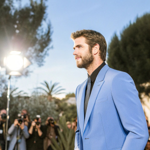 Liam Hemsworth at award show. Ruggedly handsome in navy blue suit, black shirt, and tie. Crowd of photographers and fans in background, spotlight shining on him.