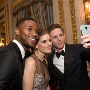 Michael B. Jordan, Kate Mara, and Jamie Bell posing for a selfie during a glamorous award show. Gilded decor and curtains in the background.