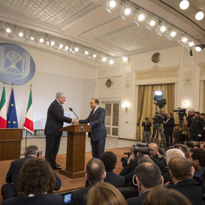 Benjamin Netanyahu and Silvio Berlusconi shaking hands at a press conference. Large room with audience and news cameras.