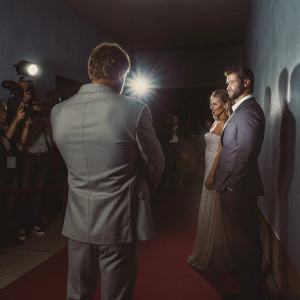 Premiere night scene. Chris Hemsworth in dark suit, Elsa Pataky in elegant gown, Kevin Feige in gray suit. Indoor event with red carpet and flashing lights.