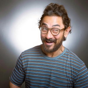 Close-up portrait of Aamir Khan. Indian actor with mustache, full beard, and warm smile. Dark, striped t-shirt, glasses with wrap-around frames, curly hair styled to the side. Solid black background.