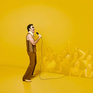 Sacha Baron Cohen as Borat, wearing a brown vest and matching pants. Performs at comedy club. Audience visible.