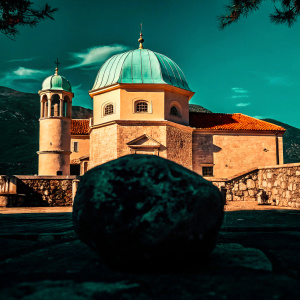An old Orthodox church located in Perast, Montenegro. The church features a striking blue dome amidst stone architecture. Set against a backdrop of clear blue skies and fluffy clouds, the scene includes a large weathered rock in the foreground and a range of mountains in the distance.