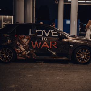 A Mitsubishi Lancer EVO X with vibrant anime-style decals and the slogan 'Love is War' in bold red and black. The car is parked outside a sleek, modern gas station with blue and gray accents. A passerby is seen in the background.