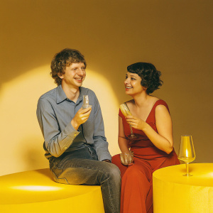 Michael Cera and Alia Shawkat smiling at a dimly lit event. Michael in a blue shirt and jeans, Alia in a red dress.