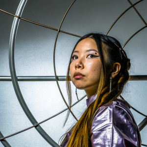 Bella Poarch, Asian model, serious face, long hair, multiple piercings, shiny purple outfit. Dark backdrop, studio photo