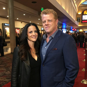 Michael and Rachel Cudlitz, both Caucasian, in a bustling lobby. Michael in a tailored navy suit, Rachel with long dark hair, at a movie premiere. Crowds in the background.