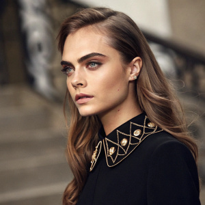 Close-up photo of Cara Delevingne. Poised look with long brown hair. Black top with intricate collar. Muted tones, blurred background