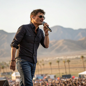 Morten Harket, singer, on stage at Coachella. Dark shirt, jeans, and shades. Surrounded by desert landscape and music festival crowd.