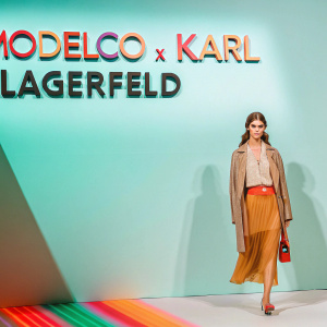A young woman stands at the Modelco x Karl Lagerfeld fashion event. A vibrant backdrop featuring the brand's logo.