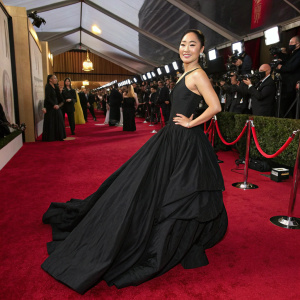 Greta Lee, an Asian actress, wearing a black, floor-length gown with a flowing skirt. She poses confidently at a awards show. Red carpet ambiance, flashing cameras.