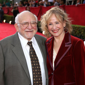 Ed Asner and Cindy Gilmore at an award ceremony. Gray suit, white shirt, patterned tie, rich burgundy velvet blazer, smiling, against red carpet backdrop.