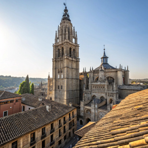 Aerial view of Toledo Cathedral from rooftop. Majestic bell tower, detailed stone work, pointed arches. Traditional Spanish buildings with terracotta roofs surround. Clear blue sky.