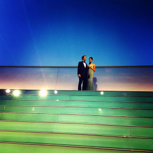 Jeremy Renner and Anna Zegna at a gala event. Both in formal attire. Brightly lit stage backdrop.