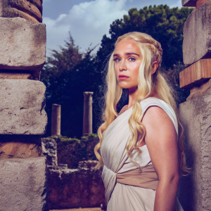 Emilia Clarke, as Daenerys Targaryen, in a stern pose. Long blonde hair, determination in eyes. Amidst ancient ruins, natural backdrop
