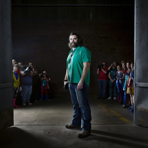 Jason Momoa in a vibrant green shirt at a comic-con event. Colorful crowd in the background.