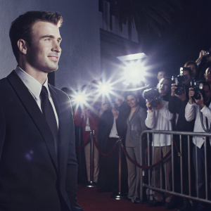 Chris Evans, in a black suit and tie, at a Hollywood premiere. Crowded and lit red carpet, paparazzi flashing lights
