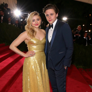 Chloe Grace Moretz and Brooklyn Beckham at the Met Gala. Chloe wears a golden sequined gown, Brooklyn in a navy blue tuxedo. They are posing on a red carpet under bright lights.