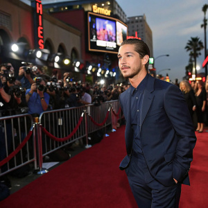 Shia LaBeouf, in a dark blue suit, stands on the bustling Hollywood red carpet. Crowds of paparazzi and fans in the background.