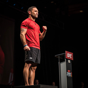 Frankie Edgar, wearing a red shirt and black shorts, standing on a UFC weigh-in platform. Dark background, showcasing his muscular physique.