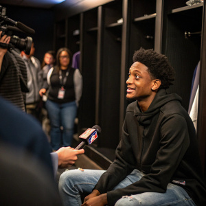 De'Aaron Fox in a locker room. Wearing a casual black hoodie and jeans. Interviews with media, blurry audience in background