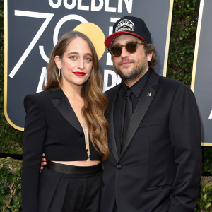 Jemima Kirke and Michael Mosberg at a chic Hollywood event. Kirke exudes confidence in a stylish ensemble, her long brown hair flowing freely. Mosberg sports a black suit, sunglasses, and a cap with a logo. The backdrop showcases the iconic Golden Globe Awards logo.
