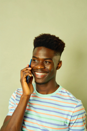 Generate an image of a young black man with a clean haircut holding a smartphone to his ear and smiling on a plain background. The camera position should be a bit far away 