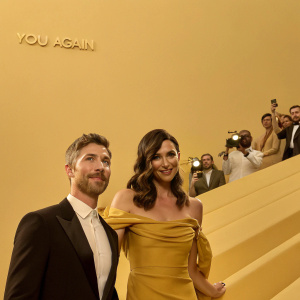 Dave Annable and Odette Yustman arriving at a bustling theater for the premiere of 'You Again'. Odette in a mustard off-the-shoulder gown, Dave in a black tuxedo. Crowds and flashing cameras in the background.