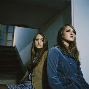Actresses Leelee Sobieski and Samantha Lang, photographed against a minimalist backdrop. Both exude elegance, with long brown hair, styled makeup, and serious expressions. Indoor setting.