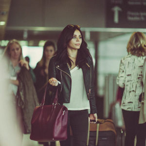 Vanessa Hudgens at a busy airport terminal. Dressed in a black leather jacket over a white shirt, black pants, and carrying a maroon handbag. Surrounded by other travelers in a dimly lit indoor setting.