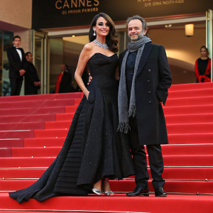 Maria Grazia Cucinotta and Giulio Violati on the Cannes Film Festival red carpet. She wears a formal black gown with silver accessories. He is in a dark coat and scarf.