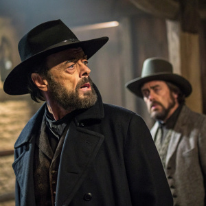 Dirk Bogarde, starring as Hermann Hermann, in a tense scene from 'Despair'. Bearded and dramatically posed. Dark-hatted actor in background. Rustic indoor setting