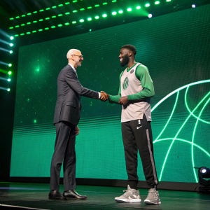 Adam Silver, NBA Commissioner, and Jaylen Brown on stage. They are shaking hands. Green LED backdrop with futuristic design.