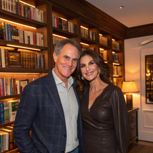 Mark Moses and Teri Hatcher at a cozy book launch party, surrounded by shelves of books and soft, ambient lighting.