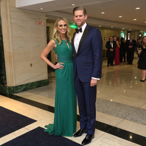 Eric Trump, in a tailored navy suit, and Lara Yunaska, in a flowing emerald green dress, are at the annual White House Correspondents' Association dinner. Black and white marble floor.
