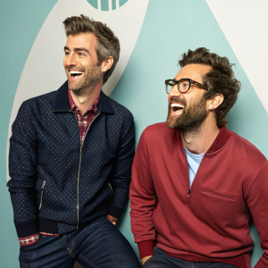 Rhett James McLaughlin and Charles Lincoln Neal III, better known as Rhett & Link, are in a jovial mood. Wearing casual attire. Backdrop features a modern graphic design.