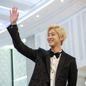 Kim Hyun Joong, Korean actor with blonde hair, waving at a press conference. Wears a black suit with a bow tie. Brightly lit, empty conference room with modern decor.